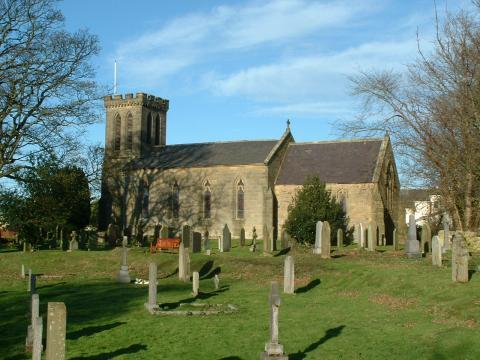 A stone church with a square tower stands in a sunlit cemetery. The church is surrounded by green grass and various headstones. Leafless trees frame the scene under a blue sky with wispy clouds.