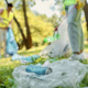 Two people wearing safety vests and gloves pick up litter in a park, collecting rubbish like cans and plastic bags from the grass into large, clear bin bags. Trees and sunlight are visible in the background.